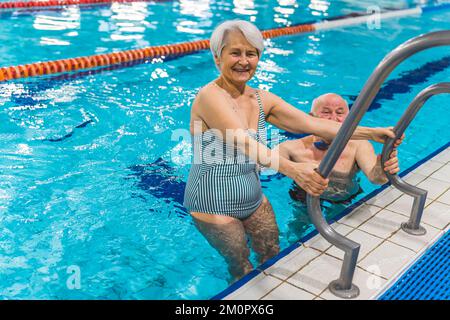 Positiv lächelnde ältere Erwachsene Frau in gestreiftem Badeanzug, die mit ihrem Mann im türkisfarbenen Becken auftaucht. Ich schaue in die Kamera. Hochwertiges Foto Stockfoto