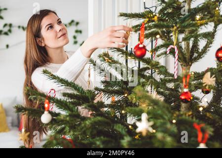 Eine lächelnde weiße Frau, die auf den natürlichen Weihnachtsbaum schaut, den sie selbst schmückt Stockfoto