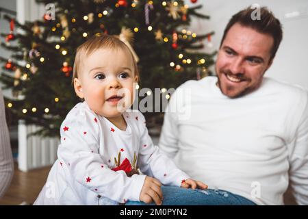 Lächelndes Mädchen, das über seinen Vater klettert, am Weihnachtstag, Weihnachtsbaum im Hintergrund Stockfoto
