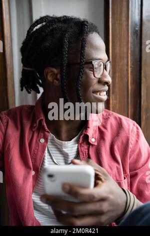 Glücklicher junger afrikanischer Mann, der auf dem Fensterbrett sitzt und ein Smartphone hält, und die mobile App für Online-Dating verwendet Stockfoto