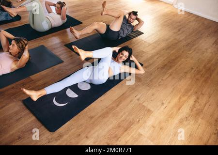 Glückliche junge Frau, die lächelt, während sie mit ihrem Kurs in einem Yoga-Studio Fahrrad-Crunches macht. Eine Gruppe sportlicher Leute, die zusammen Hatha-Yoga üben Stockfoto