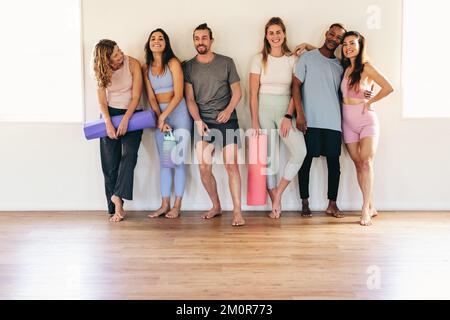 Eine Gruppe von Menschen, die mit ihren Trainingsmatten in einem Yoga-Studio lächeln und stehen. Multikulturelle Leute, die zusammen an einem Yoga-Kurs teilnehmen. Glücklich Stockfoto