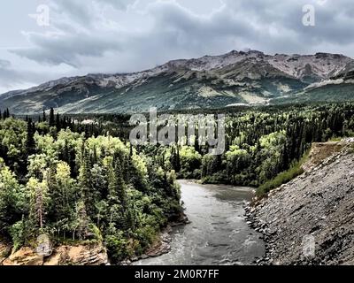 Alaska: Fluss, der durch den Wald fließt, mit einem Felsvorsprung auf der einen Seite und Bergen in der Ferne, düsterer Himmel; Fotomanipulation für Grafikeffekte Stockfoto