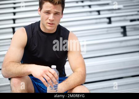 Ich habe Durst von all dem Training. Nahaufnahme des Porträts eines männlichen Läufers, der auf dem Dachsparren sitzt und eine Wasserflasche hält. Stockfoto