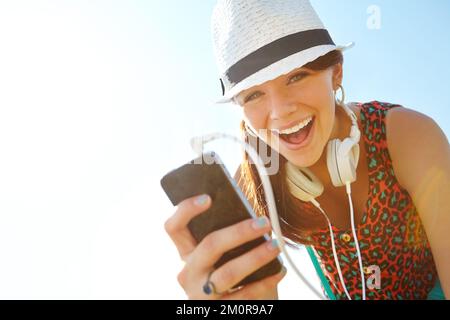 Erleben Sie den Klang des Sommers. Ein glückliches Mädchen im Teenageralter lächelt, während es ein Smartphone mit Kopfhörern hält. Stockfoto