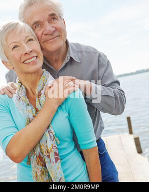 Am Seeufer zufrieden zu sein. Ein Seniorenpaar, das umarmt und nach oben schaut. Stockfoto