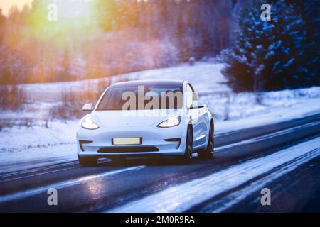 Weißes Elektroauto des Tesla Model 3 bei hoher Geschwindigkeit auf der Autobahn im Winter in der Dämmerung. Salo, Finnland. 27. Dezember 2021. Stockfoto