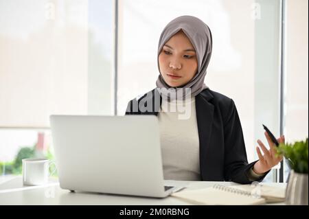 Erfolgreiche asiatische muslimische Geschäftsfrau, die Hijab trägt, einen Laptop benutzt, sich auf ihre Aufgaben konzentriert, im Büro arbeitet. Stockfoto