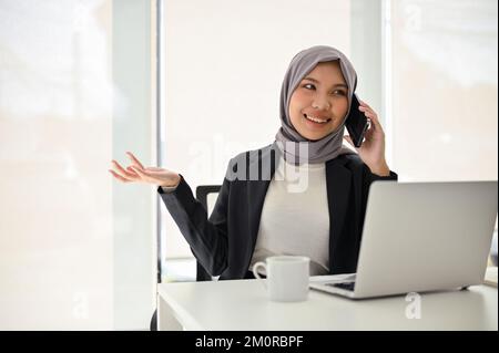 Professionelle und selbstbewusste asiatische muslimische Geschäftsfrau telefoniert mit ihrem Geschäftspartner und sitzt an ihrem Schreibtisch. Stockfoto