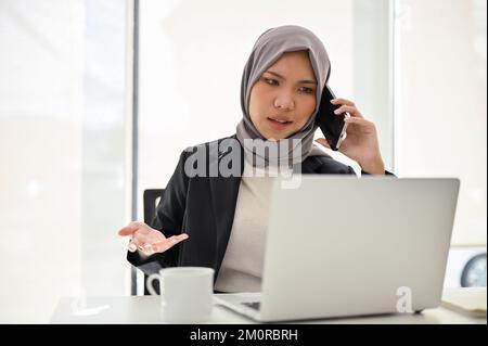 Unzufriedene und professionelle asiatische muslimische Geschäftsfrau oder weibliche Managerin, die mit dem Projektergebnis unzufrieden ist, beschwert sich telefonisch bei ihrem Mitarbeiter Stockfoto