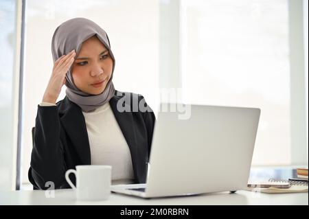 Gestresste und umsichtige asiatische muslimische Geschäftsfrau, die in ihrem Büro arbeitet, auf den Laptop-Bildschirm schaut und ihr Projekt plant. Stockfoto