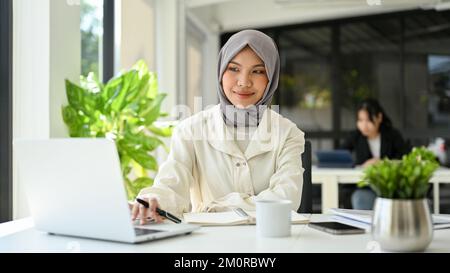 Wunderschöne, junge asiatische, muslimische Geschäftsfrau, die Hijab trägt, einen Laptop benutzt, im Büro arbeitet. Stockfoto