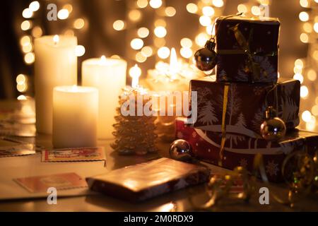 Fotoverpackte Geschenke stehen neben Kerzen auf einem Tisch mit einem Bokeh aus leuchtender Girlande im Hintergrund Stockfoto