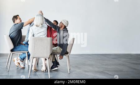 Unterschiedliche Gruppe von Menschen, die zusammensitzen und sich gegenseitig High Five geben nach Therapie mit Copyspace. Die ganze Dauer der Selbsthilfegruppe, die danach feiert Stockfoto