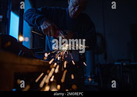 Arbeiter, Rentner mahlt Metall mit einer Schleifmaschine in seiner Werkstatt. Funken fliegen in Richtung Kamera. Stockfoto