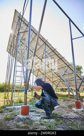 Schweißbalken mit Schweißmaschine zum Einbau von Solarpaneelen bauen. Der Mann im Schweißschutzhelm trägt Arbeitskleidung. Photovoltaiksammlung von Modulen als PV-Panel. Grüne Energie Stockfoto