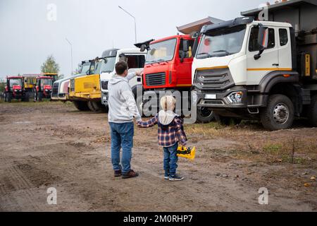 Mann und ein Junge, Vater und Sohn, stellt euch mit dem Rücken vor große Lastwagen. Die Leidenschaft des Kindes für Autos, Vater und Kind, schaut euch die Autos im Pa an Stockfoto