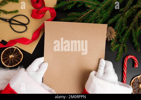 Weihnachtsmann liest Briefe von Kindern am Schreibtisch. Draufsicht auf Jolly Father Hands mit einer leeren braunen Papierseite. Blick von oben auf den Holztisch mit Stockfoto