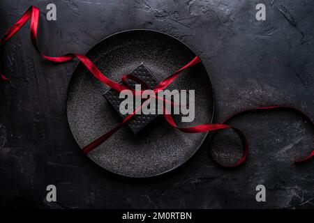 Ansicht einer schwarzen Geschenkbox mit einem roten Band auf schwarzem Schild und schwarzem Hintergrund von oben Stockfoto