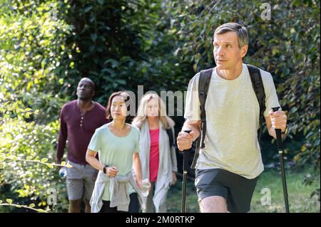 Middle-aged man carrying backpack and hiking poles while hiking in the woods with diverse group of friends Stockfoto