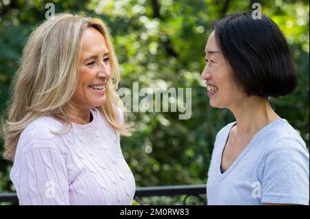 Side view shot of senior Asian-American woman and her friend smiling at each other while standing outdoors Stockfoto