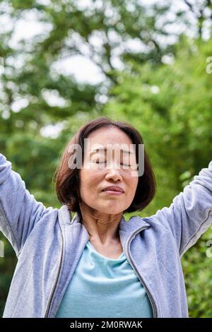 Ältere asiatische Frau, die Hände hebt, während sie tief durchatmet Stockfoto