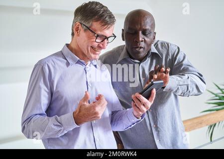 Zwei ältere, heterogene männliche Freunde, die sich zu Hause Fotos auf dem Handy ansehen Stockfoto