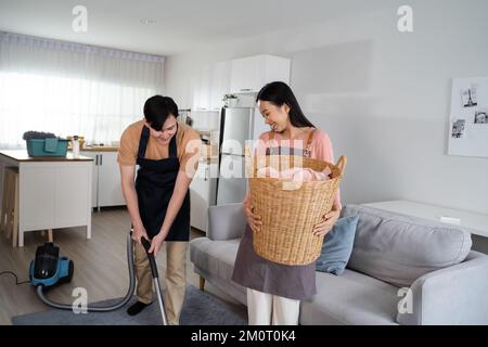 Ein junges asiatisches Paar, das zu Hause zusammen Hausbesuche macht. Eine wunderschöne Frau, die Wäsche wäscht, und ein Mann, der auf dem Boden Staubsauger macht. Hausarbeit- und Hausarbeit-Konzept Stockfoto