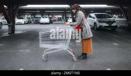 hispanische junge Erwachsene Frau, die den Wagen schiebt und die Einkaufsliste von Smartphone-Lebensmitteln auf dem Parkplatz im großen Supermarkt liest Stockfoto