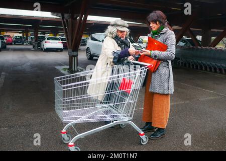 Eine europäische Seniorin und eine hispanische junge Erwachsene Frau, die einen Wagen schiebt und handgeschriebene Einkaufsliste auf dem Parkplatz eines großen Supermarkts liest Stockfoto