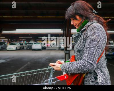 hispanische junge Erwachsene Frau, die den Wagen schiebt und die Einkaufsliste von Smartphone-Lebensmitteln auf dem Parkplatz im großen Supermarkt liest Stockfoto