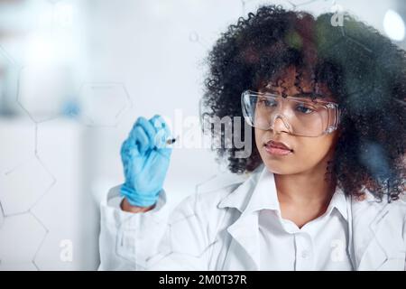 Eine ernsthafte junge Wissenschaftlerin mit einer geschwungenen Afro-Schrift und Planung auf einem Glasbrett, die bei der Arbeit Brille und Handschuhe trägt. Fokussiert Stockfoto