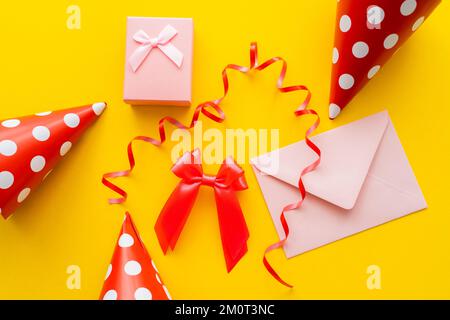 Ansicht der Geschenkbox in der Nähe des Umschlags und der Partykappen auf gelbem Hintergrund, Stockbild Stockfoto