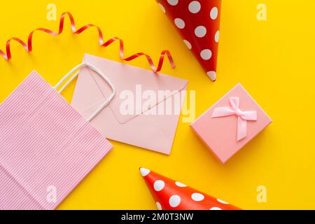 Draufsicht auf pinkfarbenen Umschlag und Einkaufstasche neben Partykappen und Geschenk auf gelbem Hintergrund, Stockbild Stockfoto