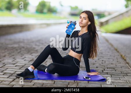 Junge schöne Frau, die einen gesunden Lebensstil führt und Musik hört, während sie auf einer Matte sitzt und sich nach Sport im Freien ausruht Stockfoto