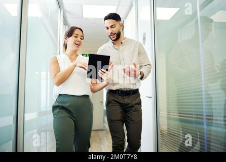 Ich brauche deine Hilfe. Zwei junge Geschäftsleute, die zusammen durch das Büro gehen und sich mit einem digitalen Tablet unterhalten. Stockfoto