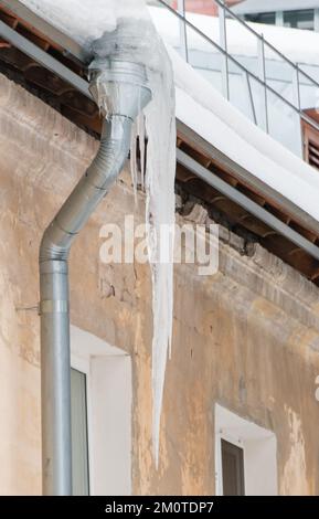 Eisige, gefrorene Eiszapfen hängen im Frühling oder Winter an einem Abflussrohr. Wand eines alten Backsteinhauses mit Fenstern. Große Eiszapfen in glatten, schönen Reihen. Bewölkter Wintertag, weiches Licht. Stockfoto
