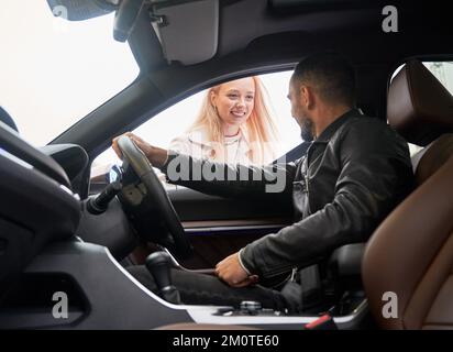 Junger, gutaussehender, glücklicher Mann, der in einem neuen Auto sitzt. Die Verkäuferin mit einem schönen Lächeln bittet darum, im Autohaus im Freien einige wichtige Dokumente zu unterschreiben. Blick aus dem Inneren eines Autos. Stockfoto