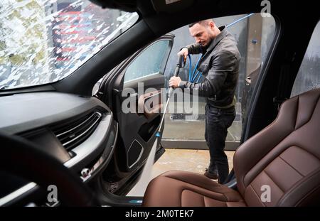 Blick vom Fahrzeuginnenraum mit öffnender Beifahrertür. Eleganter junger Mann wäscht Schmutz von der unteren Oberfläche des Autos mit Hilfe eines Spezialwerkzeugs ab, das Hochdruckwasser liefert. Stockfoto