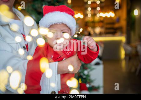 Süßes neugeborenes Kind mit Weihnachtsmannmütze und festlichem Outfit neben einem Weihnachtsbaum mit festlicher Dekoration und Fährenbeleuchtung für Neujahr und Weihnachten Stockfoto