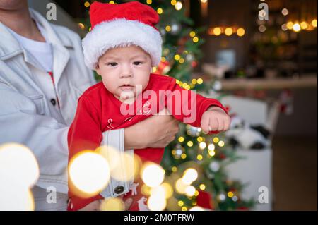 Süßes neugeborenes Kind mit Weihnachtsmannmütze und festlichem Outfit neben einem Weihnachtsbaum mit festlicher Dekoration und Fährenbeleuchtung für Neujahr und Weihnachten Stockfoto