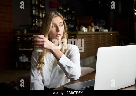 Porträt einer jungen, hübschen blonden Frau, die an einem Tisch in einem schwarzen Stuhl sitzt, an einem Laptop arbeitet und ein Glas Cappuccino teilt Stockfoto