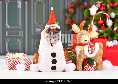 Lustiger französischer Bulldog in Weihnachtskostüm und Weihnachtsmannmütze vor saisonaler Dekoration Stockfoto