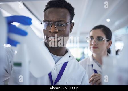 Junge afroamerikanische Wissenschaftlerin, die die Probenflasche mit einer Kollegin überprüft.zwei Mediziner, die gemeinsam an Experimenten im Labor arbeiten Stockfoto
