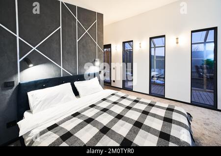 Modernes Schlafzimmer mit zahlreichen Glastüren, die zur Terrasse in der Abenddämmerung führen. Seitenansicht des beleuchteten großen Betts mit Karottendecke vor der grauen Wand im Schlafbereich im Innenbereich. Innenkonzept. Stockfoto