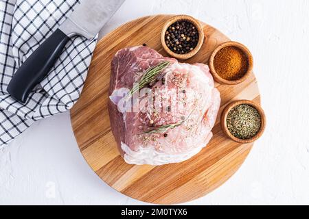 Ein großes frisches Stück Schweinefleisch auf einem runden Holzbrett mit Schüsseln verschiedener Gewürze zur Zubereitung der Marinade. Draufsicht. Weißer Hintergrund Stockfoto