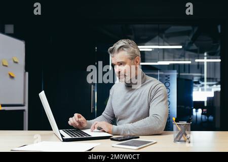 Seriöser und behutsamer, reifer Investor, der in modernen Büros arbeitet, leitender, grauhaariger Geschäftsmann, der bei der Arbeit ein Notebook benutzt, männlicher Boss, der am Schreibtisch sitzt. Stockfoto