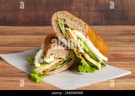 Baguette-Sandwich. Sandwich mit Oliven, Salat, Tomaten, Gurken, Cheddar und Streichkäse auf Holzhintergrund Stockfoto