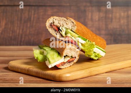 Baguette-Sandwich. Sandwich mit Oliven, Salat, Tomaten, Gurken, Cheddar und Fetakäse auf Holzhintergrund Stockfoto