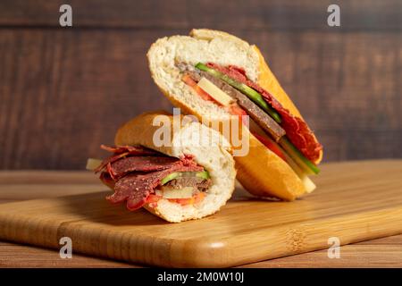 Baguette-Sandwich. Sandwich mit Salami, Roastbeef, Cheddar, Tomaten und Gurke auf Holzhintergrund Stockfoto